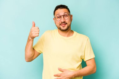 Mavi arka planda izole edilmiş genç İspanyol adam karnına dokunuyor, nazikçe gülümsüyor, yemek yiyor ve tatmin oluyor..