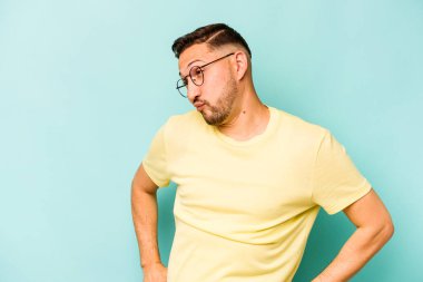 Mavi arka planda izole edilmiş genç bir İspanyol omuz silker ve gözleri açık kalır..