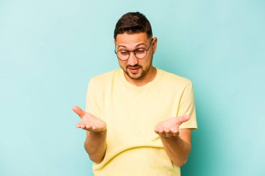 Genç İspanyol bir adam mavi arka planda izole edilmiş. Elinde avuç içi olan bir şeyle kameraya sunuyor..