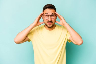 Genç İspanyol bir adam mavi arka planda izole edilmiş. Hoş bir sürpriz bekliyor. Heyecanlı ve el kaldırıyor..