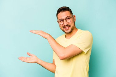 Mavi arka planda izole edilmiş genç İspanyol adam işaret parmaklarıyla küçük bir şey tutuyor, gülümsüyor ve kendinden emin..