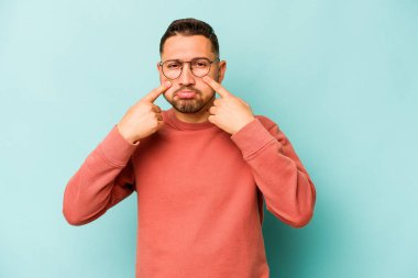 Genç İspanyol bir adam mavi arka planda izole edilmiş, ağlıyor, bir şeyden memnun değil, acı ve kafa karışıklığı konsepti..