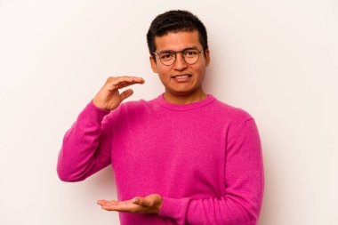 Beyaz arka planda izole edilmiş genç İspanyol adam işaret parmaklarıyla küçük bir şey tutuyor, gülümsüyor ve kendinden emin..