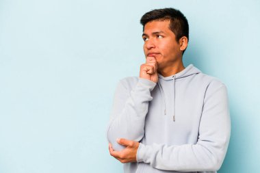 Genç İspanyol adam mavi arka planda izole edilmiş şüpheli ve şüpheci bir ifadeyle yana bakıyor..