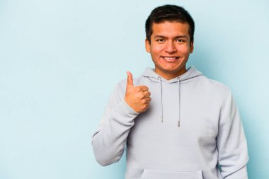 Genç İspanyol adam mavi arka planda izole edilmiş gülümsüyor ve başparmağını kaldırıyor.