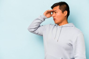 Genç İspanyol adam mavi arka planda izole edilmiş, uzaklara bakıyor ve elini alnına koyuyor..