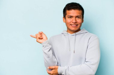 Mavi arka planda izole edilmiş genç İspanyol adam işaret parmağıyla işaret ederek gülümsüyor..