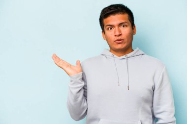 Mavi arka planda izole edilmiş genç İspanyol adam, avuç içinde kopya alanı tutmaktan etkilenmiş..