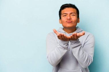 Genç İspanyol adam mavi arka planda izole edilmiş dudaklar kıvrılıyor ve hava öpücüğü göndermek için avuç içleri tutuyor..