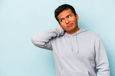 Genç İspanyol bir adam mavi arka planda izole edilmiş kafasının arkasına dokunuyor, düşünüyor ve bir seçim yapıyor..