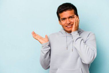 Mavi arka planda izole edilmiş genç İspanyol bir adam avucunun içinde fotokopi alanı tutuyor, el üstünde tutuyor. Şaşırmış ve mutlu..