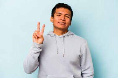Mavi arka planda izole edilmiş genç İspanyol adam zafer işareti gösteriyor ve gülümsüyor..