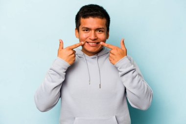 Mavi arka planda izole edilmiş genç İspanyol adam gülümsüyor, parmaklarını ağıza doğrultuyor..