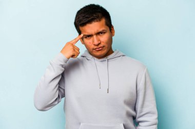 Genç İspanyol bir adam mavi arka planda izole edilmiş, parmağıyla tapınağı işaret ediyor, düşünüyor ve bir göreve odaklanıyor..