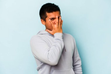 Mavi arka planda izole edilmiş genç İspanyol adam kameraya parmaklarıyla göz kırpıyor, yüzünü kapatmaktan utanıyor..