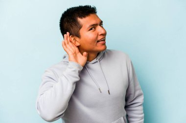 Genç İspanyol bir adam mavi arka planda izole edilmiş bir şekilde bir dedikoduyu dinlemeye çalışıyor..