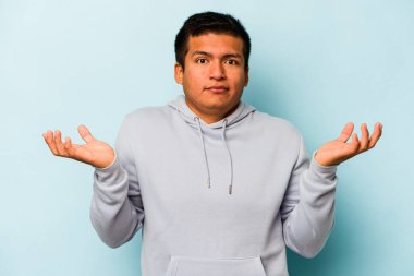 Latin kökenli genç bir adam, mavi arka planda izole edilmiş şüphe içinde ve sorgulama sırasında omuz silkiyor..
