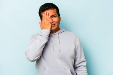 Genç İspanyol adam mavi arka planda izole edilmiş. Yüzünün yarısını avuç içiyle kaplayarak eğleniyor..
