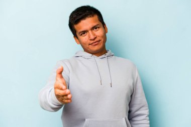 Mavi arka planda izole edilmiş genç İspanyol adam selamlama jesti olarak kameraya uzanıyor..