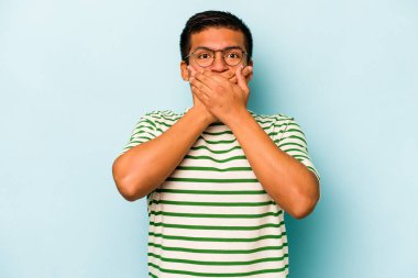 Mavi arka planda izole edilmiş genç İspanyol adam elleriyle ağzını kapatırken şok oldu..