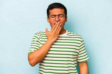 Mavi arka planda izole edilmiş genç İspanyol adam esniyor. Ağzı eliyle kapatan yorgun bir hareket sergiliyor..