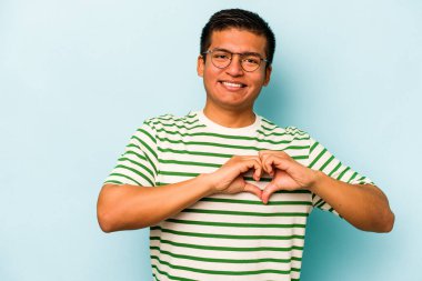 Mavi arka planda izole edilmiş genç İspanyol adam gülümsüyor ve elleriyle kalp şeklini gösteriyor..