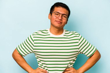 Latin kökenli genç adam mavi arka planda izole edilmiş. Kendine güveniyor. Kalçaları tutuyor..