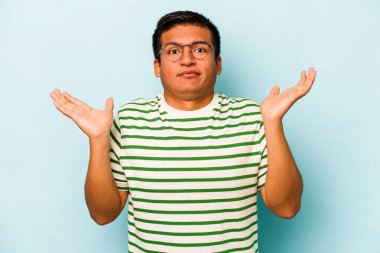 Genç İspanyol bir adam mavi arka planda izole edilmiş. Kafası karışmış ve omuz silkiyor. Kopya alanını tutmak için..