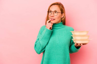 Beyaz tenli genç bir kadın pembe arka planda izole edilmiş tupperware taşıyor. Bir şeyin fotokopi odasına baktığını düşünüyor..