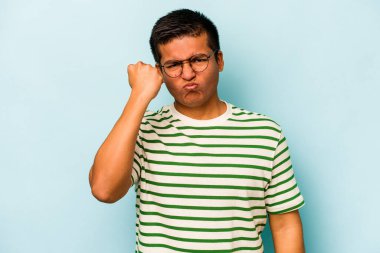 Mavi arka planda izole edilmiş genç İspanyol adam kameraya yumruk gösteriyor, agresif yüz ifadesi..