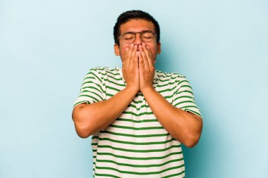 Genç İspanyol bir adam mavi arka planda izole edilmiş bir şekilde bir şeye gülüyor, elleriyle ağzını kapatıyor..