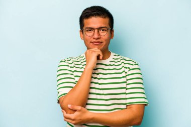 Genç İspanyol adam mavi arka planda izole edilmiş mutlu ve kendinden emin gülümsüyor, elleriyle çenesine dokunuyor..