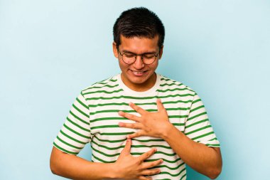 Mavi arka planda izole edilmiş genç İspanyol adam mutlu bir şekilde gülüyor ve ellerini karnının üstünde tutarak eğleniyor..