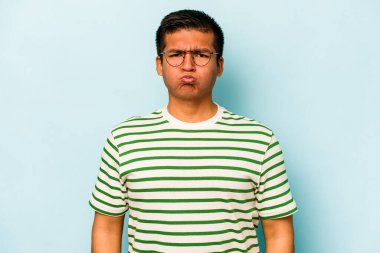 Mavi arka planda izole edilmiş genç İspanyol adam yanakları patlatıyor, yorgun bir ifadesi var. Yüz ifadesi kavramı.