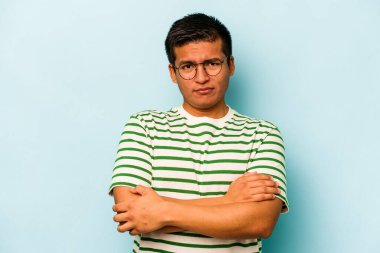 Latin asıllı genç adam mavi arka planda izole edilmiş. Mutsuz bir şekilde kameraya bakıyor. İğneleyici bir ifadeyle..