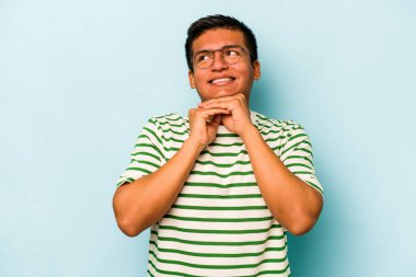 Mavi arka planda izole edilmiş genç İspanyol adam ellerini çenesinin altında tutuyor ve mutlu bir şekilde yan tarafa bakıyor..