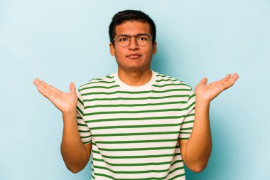 Mavi arka planda izole edilmiş genç İspanyol adam kollarıyla ölçeği yapıyor, mutlu ve kendinden emin hissediyor..