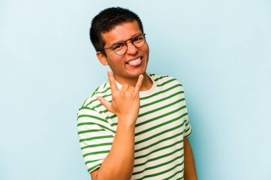 Mavi arka planda izole edilmiş genç İspanyol adam parmaklarıyla kaya hareketi gösteriyor.