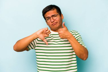Latin kökenli genç adam mavi arka planda izole edilmiş, baş parmağını kaldırıyor, zor bir seçim konsepti.