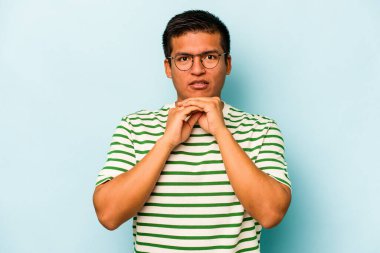 Latin kökenli genç adam mavi arka planda izole edilmiş şans için dua ediyor, şaşırmış ve ağzı açık bir şekilde önüne bakıyor..