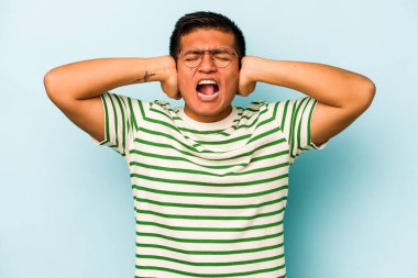 Genç İspanyol bir adam mavi arka planda izole edilmiş kulaklarını elleriyle kapatıyor çok fazla ses duymamaya çalışıyor..