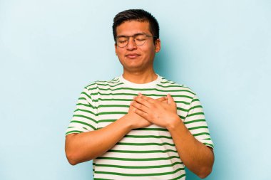 Mavi arka planda izole olmuş genç İspanyol adam gülüyor, ellerini yüreğine koyuyor, mutluluk kavramını koruyor..