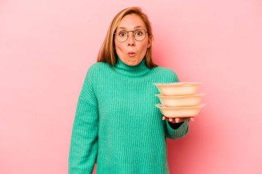 Beyaz tenli genç bir kadın pembe arka planda izole edilmiş tupperware taşıyor omuzlarını silkiyor ve gözleri açık..