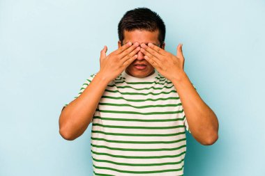Mavi arka planda izole edilmiş genç İspanyol adam elleriyle gözlerini kapatmaktan korkuyor..