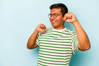 Genç İspanyol adam mavi arka planda izole edilmiş özel bir günü kutluyor, atlıyor ve enerjiyle kollarını kaldırıyor..