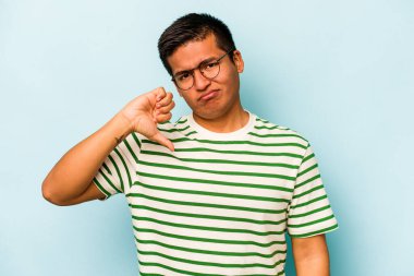 Mavi arka planda izole edilmiş genç İspanyol adam baş parmağını aşağı gösteriyor, hayal kırıklığı konsepti.