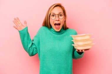 Beyaz tenli genç bir kadın pembe arka planda saklanmış vaziyette güzel bir sürpriz bekliyor, heyecanlı ve el kaldırıyor..