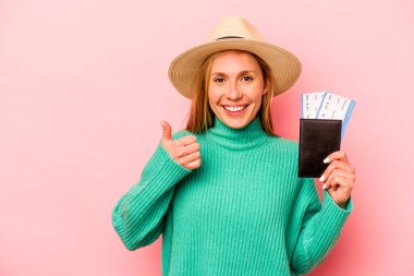Pasaportu olan beyaz bir kadın pembe arka planda izole edilmiş gülümsüyor ve başparmağını kaldırıyor.