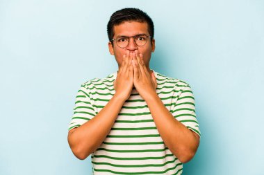 Mavi arka planda izole edilmiş genç İspanyol adam şok olmuş, ağzı ellerle kaplanmış, yeni bir şey keşfetmek için sabırsızlanıyor..