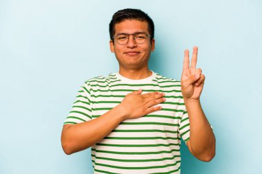 Genç İspanyol bir adam mavi arka planda izole edilmiş yemin ediyor, elini göğsüne koyuyor..